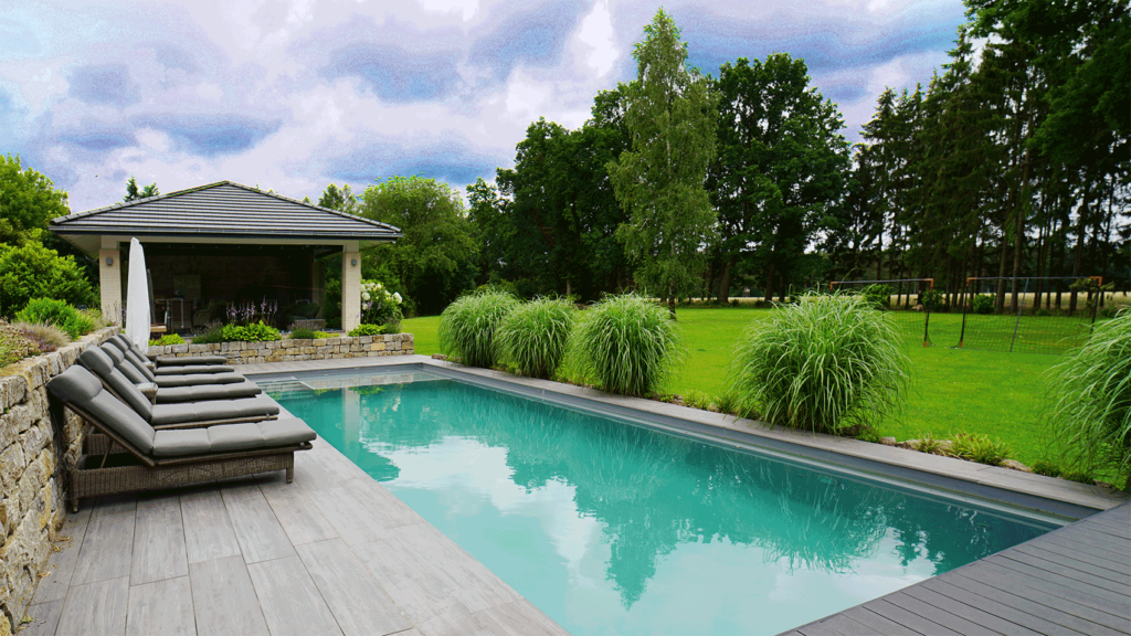 Großer Garten mit langem Pool. Grüne Gräser und Relax-Liegen umzäunen den Pool.