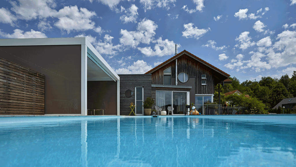 Blick zum Wohnhaus mit blauem Wolkenhimmel. Im Vordergrund der Pool mit türkisfarbenem Wasser.