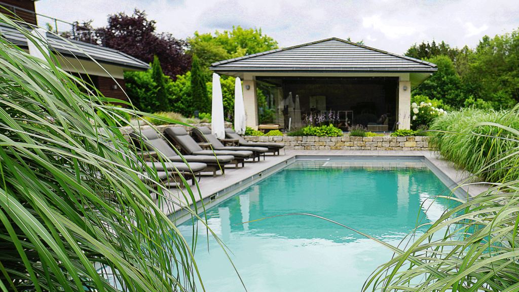 Blick durch die grüne Bepflanzung auf den Pool und das Gartenhaus im Hintergrund. Der Pool ist Ringsum üppig bepflanzt.