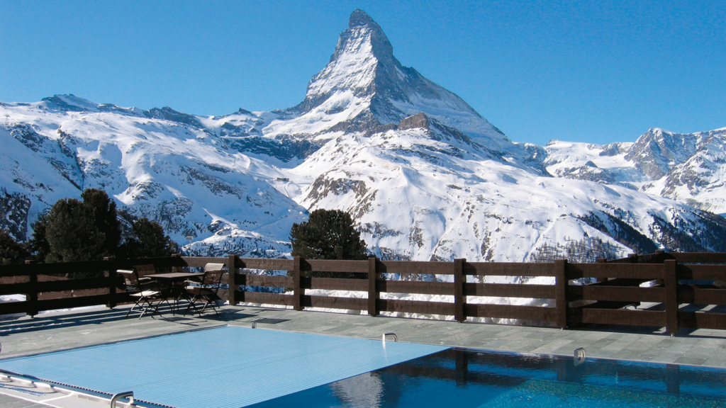Im Hintergrund ein Bergpanorama der Schweiz mit dem Matterhorn im Zentrum, im Vordergrund eine Terrasse und ein großer Pool, dessen Rollladenabdeckung zur Hälfte geschlossen ist.