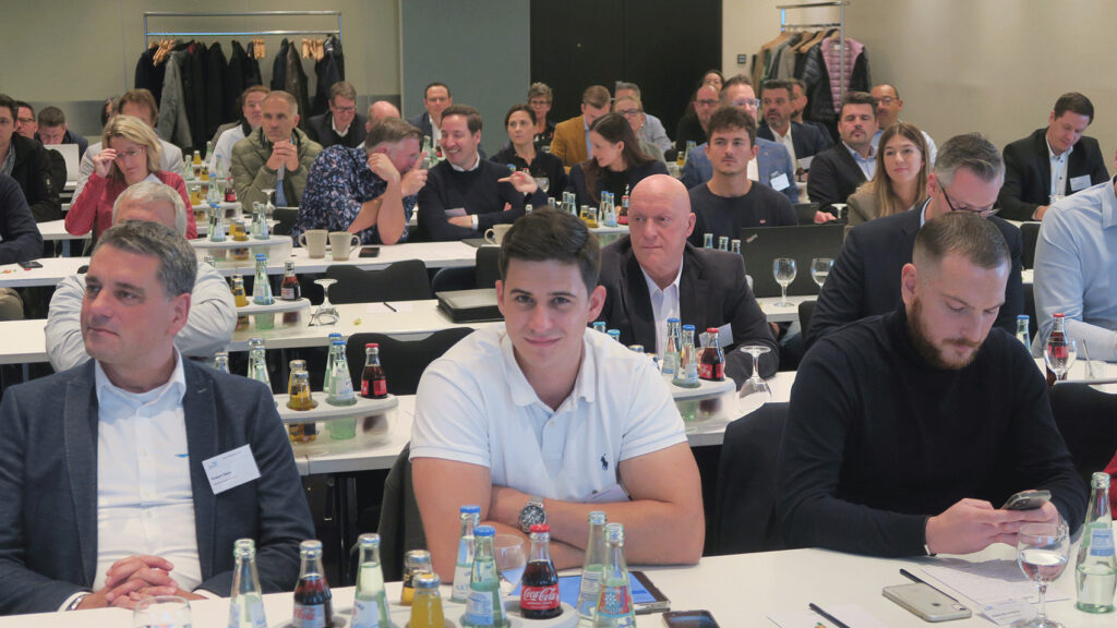 Blick in einen gut gefüllten Konferenzraum. Die Menschen sitzen in Reihen hintereinander und lauschen einem Vortrag, auf den Tischen stehen Getränke und es liegen Notizen vor den Teilnehmern.