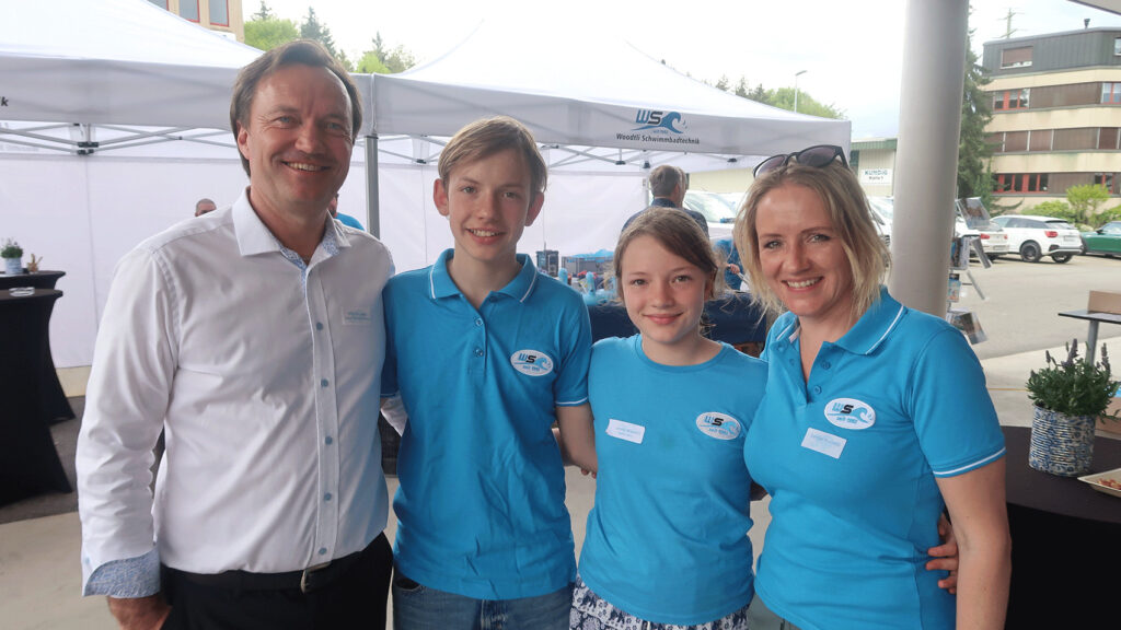 Eltern posieren mit ihren beiden Kindern in blauen -Shirts mit Firmenlogo bei einem Event auf dem Außengelände einer Firma.