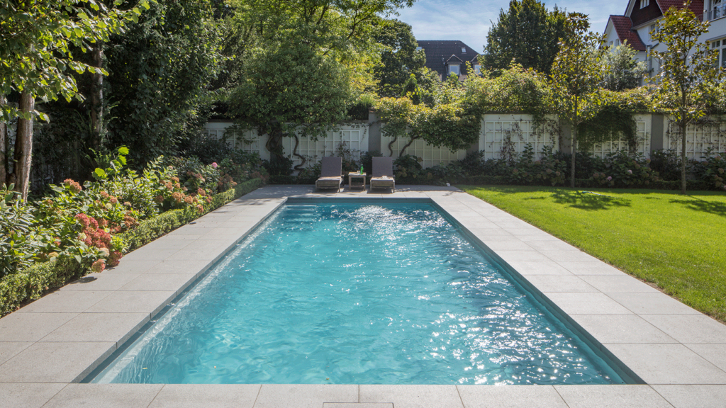 Fertigschwimmbecken mit Gegenstromanlage in einem Garten von gefliester Terrasse umgeben.