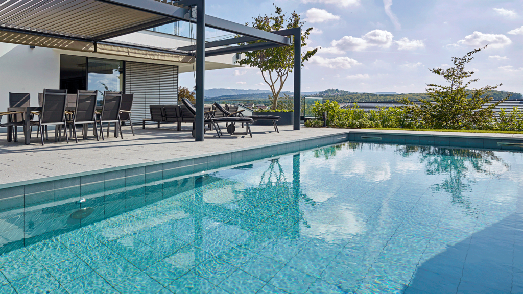 Im Vordergrund ein gefliester Pool, dahinter die Terrasse und ein modernes Wohnhaus sowie der Blick in die weite Landschaft.