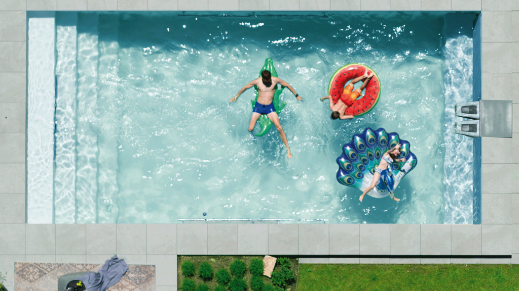 Ein Pool von oben, auf dem Wasser schwimmend drei Jugendliche auf aufblasbaren Wassertieren. Rechts im Bild ist eine Turbinenschwimmanlage im Pool zu sehen.