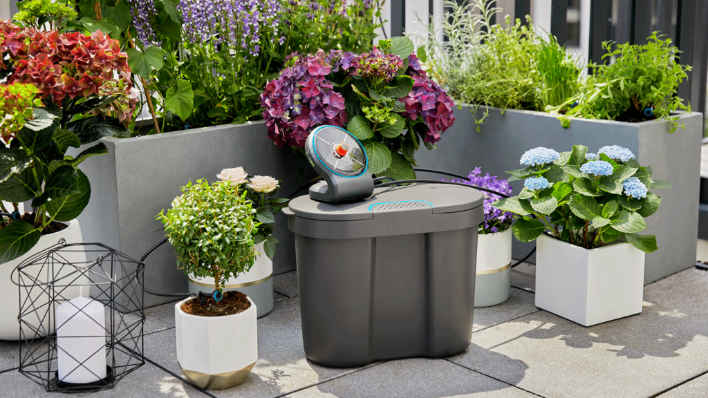 Der Wassertank und der Solarkollektor des Gardena Aquabloom stehen auf einem Balkon vor bunten Hortensien in Töpfen verschiedener Größen.