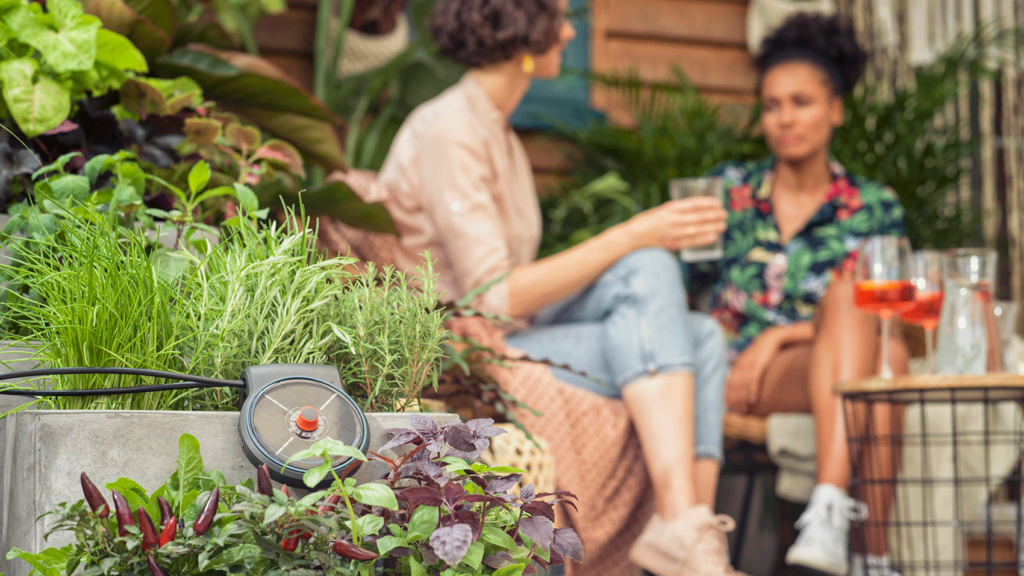 Im Vordergrund bepflanzte Blumenkästen mit einem Gardena-Solarkollektor, im Hintergrund zwei Frauen, die bei kühlen Getränken auf der Terrasse entspannen.