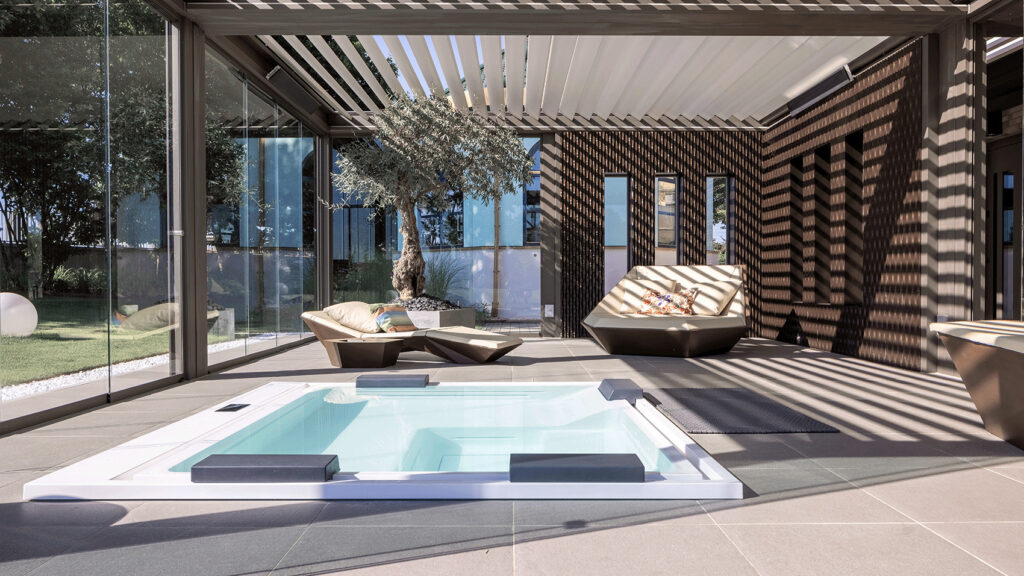 Eine Terrasse mit in die Erde eingelassenem Whirlpool und Lounge-Sitzmöbeln. Überdacht von einem Lamellendach und mit Glaswänden.