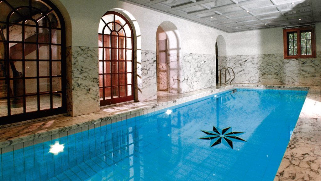 Eine mit Marmor ausgekleidete Schwimmhalle mit gefliestem Pool und bodentiefen Rundbogenfenstern sowie mit Säulengang