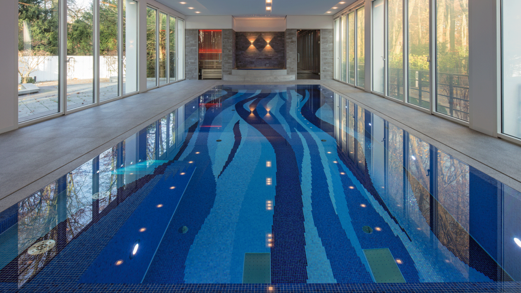 Eine Schwimmhalle mit einem Pool, der mit Mosaiksteinchen ausgekleidet ist. Die Mosaike bilden Wellenmuster und sind in verschiedenen Blautönen gehalten.