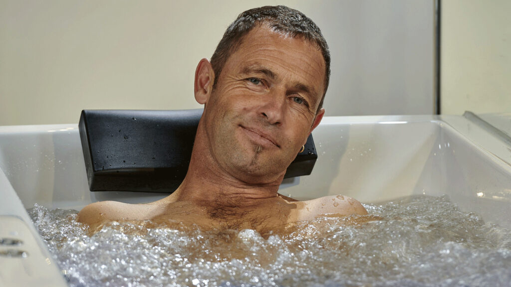 Ein Mann lächelt und sitzt in einer Badewanne mit Massagedüsen in sprudelndem Wasser.