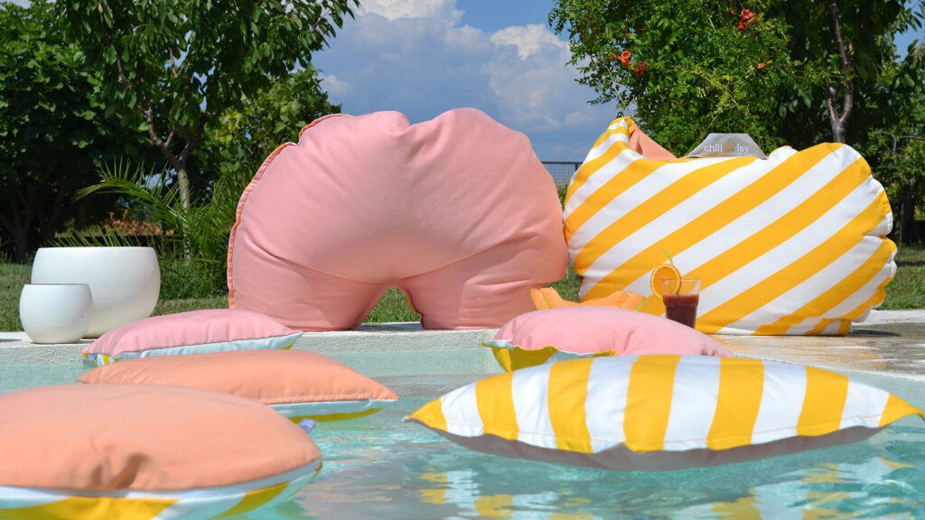 Mehrere farbenfrohe schwimmende Kissen in apricot und weiß/gelb-gestreift schwimmen auf der Wasseroberfläche eines Pools und weitere Kissen liegen auf der angrenzenden Terrasse.