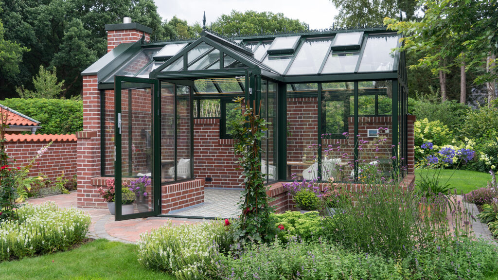 Ein großes Gewächshaus aus rotem Backstein, schwwarzem Gusseisen und viel Glasfläche in einem Bauerngarten.