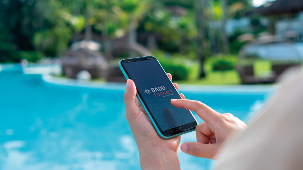 Blick auf ein Smartphone-Display auf dem die Poolsteuerungs-App BADU Connect zu sehen ist, im Hintergrund ein Pool.