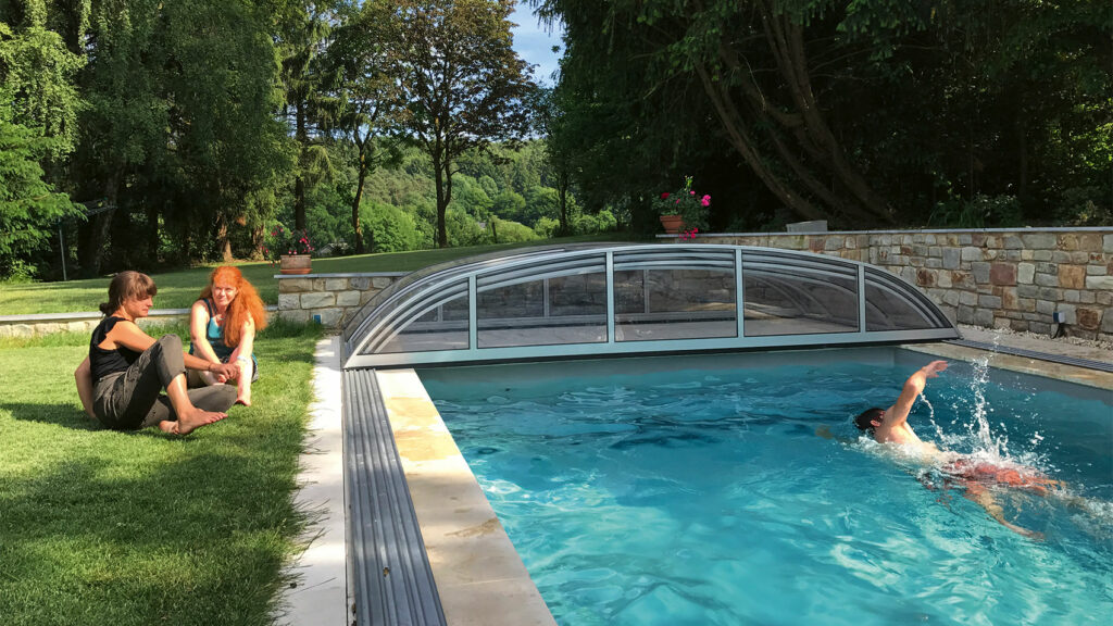 Garten mit vielen Bäumen, Rasenflächen und einem Pool mit zusammengeschobener Überdachung. Am Rand sitzen zwei Frauen. Im Pool schwimmt ein Mann.