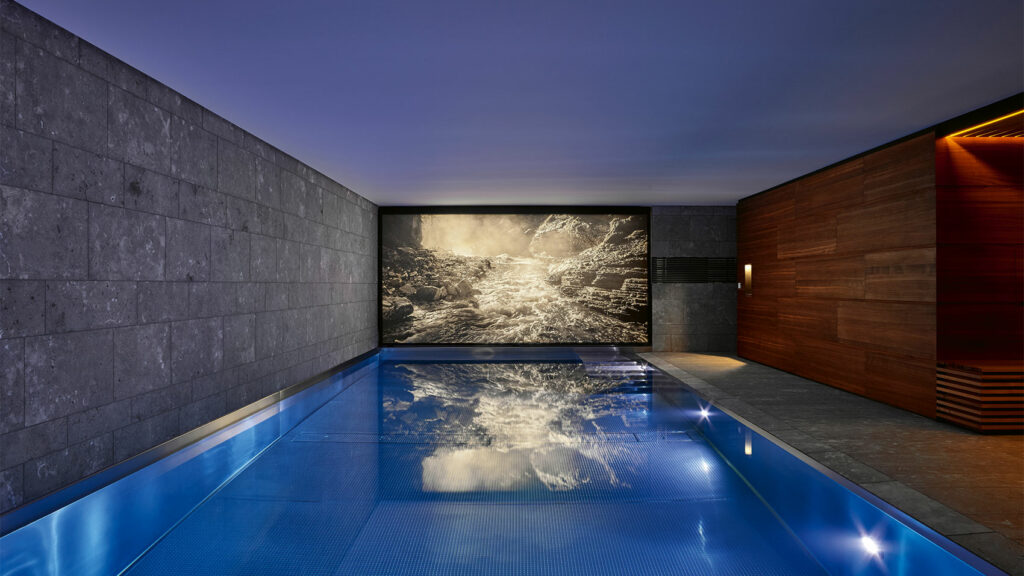 Schwimmhalle mit Edelstahlpool und einem Naturbild an der Stirnseite, welches eine von hinten beleuchtete Flusslandschaft zeigt.