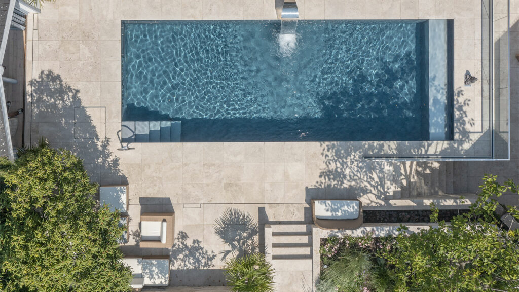 Blick von oben auf einen Pool mit Schwalldusche eingebettet in eine Terrasse aus hellen Travertinsteinen.