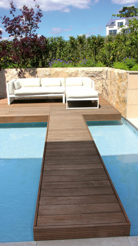 Holzsteg, der über das Wasser eines Pools zu einem Holzdeck mit gemütlicher Gartenlounge führt.